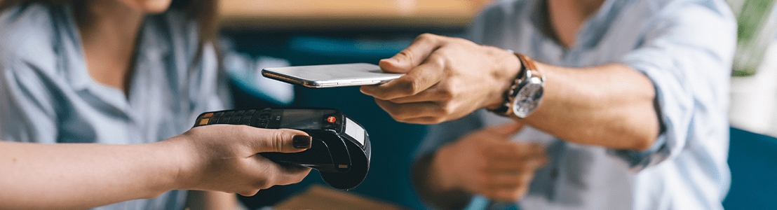 Im Vordergrund ist hält eine Hand ein mobiles EC-Terminal während eine andere Hand mit einer EC-Karte an diesem Terminal bezahlt. Im Hintergrund sind die Umrisse eines Mannes und einer Frau zu sehen.