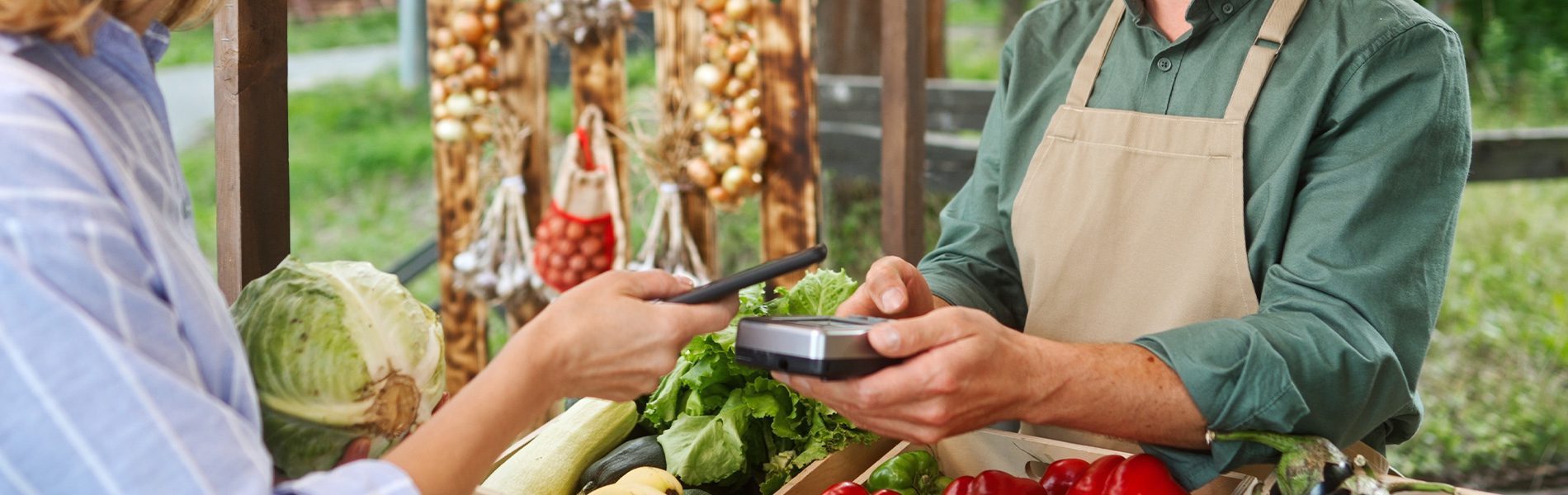 Kontaktlose Kartenzahlung im Einzelhandel: Kundin zahlt mit Smartphone an einem mobilen EC-Terminal am Gemüsestand eines Händlers mit Schürze.