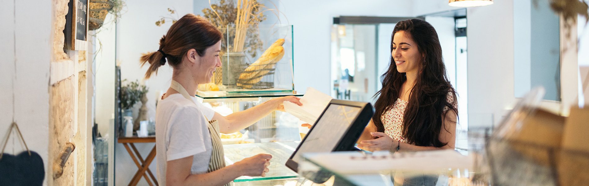 Bäckerei-Tresen, an dem eine Mitarbeiterin eine Backwaren-Tüte an Kundin übergibt; modernes EC-Terminal/Kassensystem neben Glasvitrine mit Baguettes – Gastronomie.