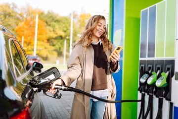 Person tankt Auto an moderner Tankstelle und hält ein Smartphone; Zapfsäule mit grünen Kraftstoffdüsen im Hintergrund.