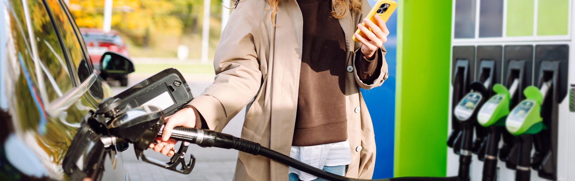 Person tankt Auto an moderner Tankstelle und hält ein Smartphone; Zapfsäule mit grünen Kraftstoffdüsen im Hintergrund.